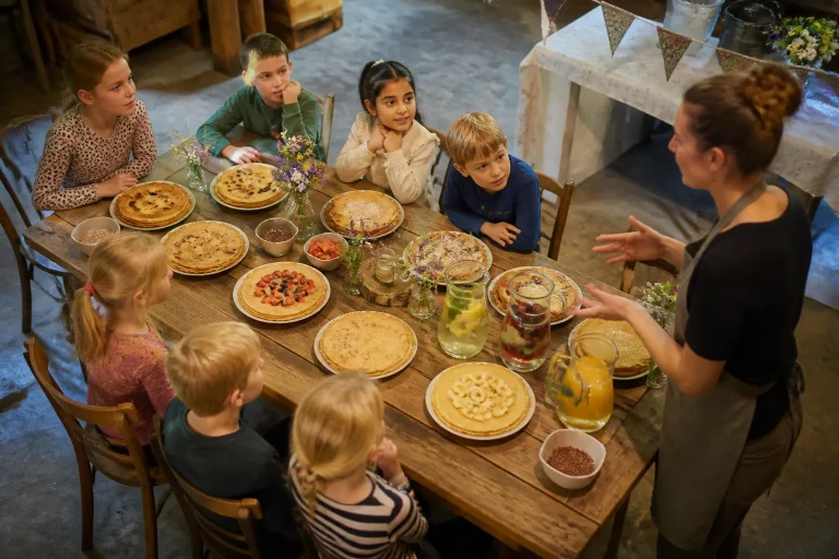 kinderen rond lange houten boerderijtabel vol pannenkoeken terwijl volwassene tips geeft